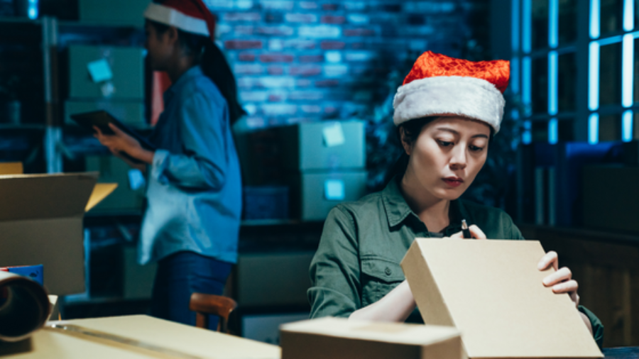 Two women in warehouse wearing santa hats, packing online orders for shipment.